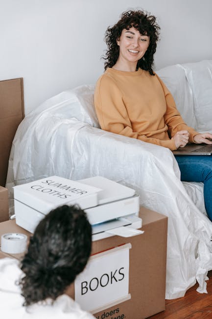 Image showing a young woman with curly dark hair sitting on a white sofa in a bright room, smiling and using a laptop. In front of her, there are several cardboard boxes, one labeled 'BOOKS' and anoth