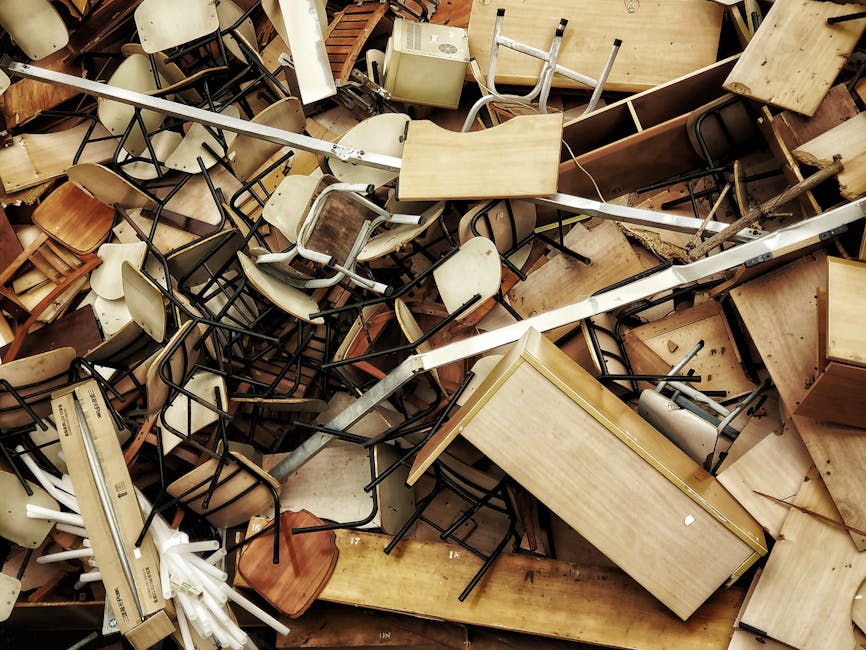 A large pile of discarded furniture and household items, including various wooden desks, chairs, and tables, along with some office equipment and packaging materials such as cardboard and plastic wrap