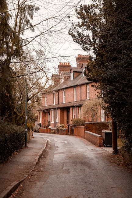 A narrow residential street lined with brick houses, with some featuring small front gardens and low brick walls. The street appears to be in a quiet neighbourhood with bare trees and overcast sky, indicating a late autumn or winter setting. The road surface is paved, with a slight bend visible ahead. The environment suggests a typical suburban environment suitable for house removals and home relocation services. The scene is captured during daylight hours with natural, subdued lighting and no moving vehicles or personnel visible, providing a neutral backdrop for discussing packing, furniture transport, and loading processes by companies like Man with Van Surbiton as part of their house removals in Surbiton.