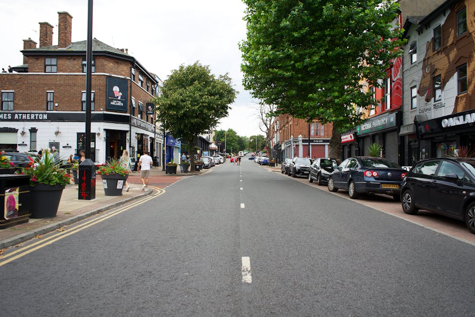 The image shows a busy street in Surbiton with a row of traditional brick and white-painted terraced houses facing the pavement. Some of the buildings have shopfronts, including a bakery and a café, with signs visible for customers. In front of the shops, several cars are parked along the curb, including a black car, a white car, and a silver vehicle. The street features two red double-decker buses, indicating public transport access, and a few pedestrians are walking along the sidewalk. On the right side of the image, a large building is under construction, covered in white scaffolding and plastic sheeting. The scene is captured during daylight hours, with overcast weather, and the overall setting reflects a typical urban environment where house removals and furniture transport services by [COMPANY_NAME] like Man with Van Surbiton could be required for efficient home relocation and moving logistics.