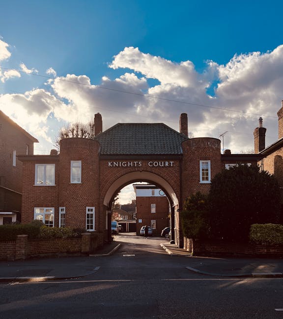 Photograph of Knights Court, a brick residential building with an arched entrance, situated on Kingston Road in Surbiton. The building features chimneys on the roof, multiple windows, and a small brick wall with shrubbery on either side of the entrance. Inside the archway, a parking area is visible with several cars parked, and part of a van is seen nearby, indicating movement of household items. The scene is lit by natural daylight with a partly cloudy sky overhead. This image relates to house removals and furniture transport, illustrating the typical environment where professional moving services, such as those offered by Man with Van Surbiton, operate during home relocations involving loading and unloading processes outside residential properties.