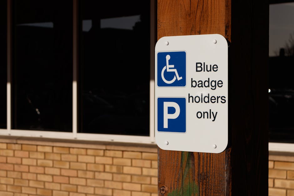 A close-up photograph of a white rectangular parking sign mounted on a wooden post outside a building with large glass windows and a brick facade. The sign displays a blue wheelchair symbol indicating accessibility, along with the text 'Blue badge holders only,' and a blue 'P' symbol, signifying designated parking for disabled badge holders. The sign is positioned on the pavement next to the building, suggesting it delineates parking restrictions for accessible vehicles during a home relocation or furniture transport process. The background shows reflections in the windows, indicating an urban setting, with lighting that appears to be natural daylight. This image relates to parking permit requirements for removals, as managed by [COMPANY_NAME], and is relevant within the context of house removals and moving logistics on Kingston Road, Surbiton.
