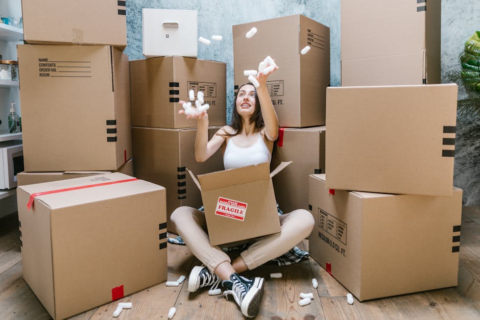 A young woman with light skin and long dark hair sits cross-legged on a wooden floor amid numerous cardboard moving boxes in a room with a textured blue wall and a white desk with a bottle of hand sanitizer. She is wearing a white tank top, light-colored pants, and black-and-white sneakers. She is smiling and looking upwards as she tosses white packing peanuts into the air, with some falling into the open flaps of a medium-sized cardboard box in her lap labeled 'Fragile'. The boxes around her vary in size, some sealed with red tape, others open or partially filled. Behind her, a tall stack of boxes is placed against the wall, and nearby, a green potted plant is partially visible. The scene depicts the process of packing and preparing for a house move, representative of packing and moving logistics carried out by Man with Van Surbiton, a professional removals service specializing in home relocation and furniture transport in Surbiton.