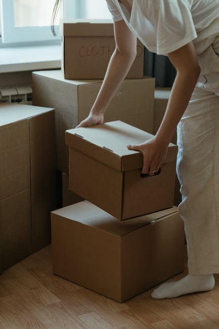 Inside a residential property, a person wearing a beige shirt and white trousers is seen lifting and stacking several cardboard boxes used for packing household items, including clothing and personal belongings. The boxes are of various sizes, some labeled with handwritten text such as 'CLOTH'. The individual is standing on a wooden floor near a window that provides natural daylight. Behind them, there are additional wrapped and unpacked boxes arranged for a home relocation process. The scene depicts the packing phase of a house move managed by Man with Van Surbiton, focusing on the careful handling of packed contents in preparation for furniture transport and removal services. The environment suggests an organized approach to packing and loading, with items ready for moving between locations as part of the home removals process.