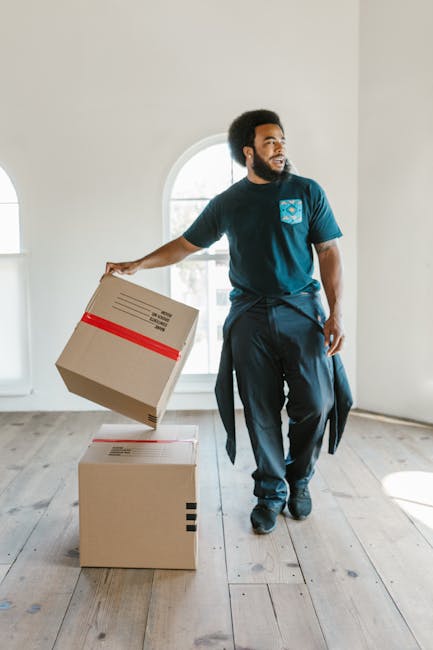 A young woman with light skin and long dark hair sits cross-legged on a wooden floor amid numerous cardboard moving boxes in a room with a textured blue wall and a white desk with a bottle of hand sanitizer. She is wearing a white tank top, light-colored pants, and black-and-white sneakers. She is smiling and looking upwards as she tosses white packing peanuts into the air, with some falling into the open flaps of a medium-sized cardboard box in her lap labeled 'Fragile'. The boxes around her vary in size, some sealed with red tape, others open or partially filled. Behind her, a tall stack of boxes is placed against the wall, and nearby, a green potted plant is partially visible. The scene depicts the process of packing and preparing for a house move, representative of packing and moving logistics carried out by Man with Van Surbiton, a professional removals service specializing in home relocation and furniture transport in Surbiton.