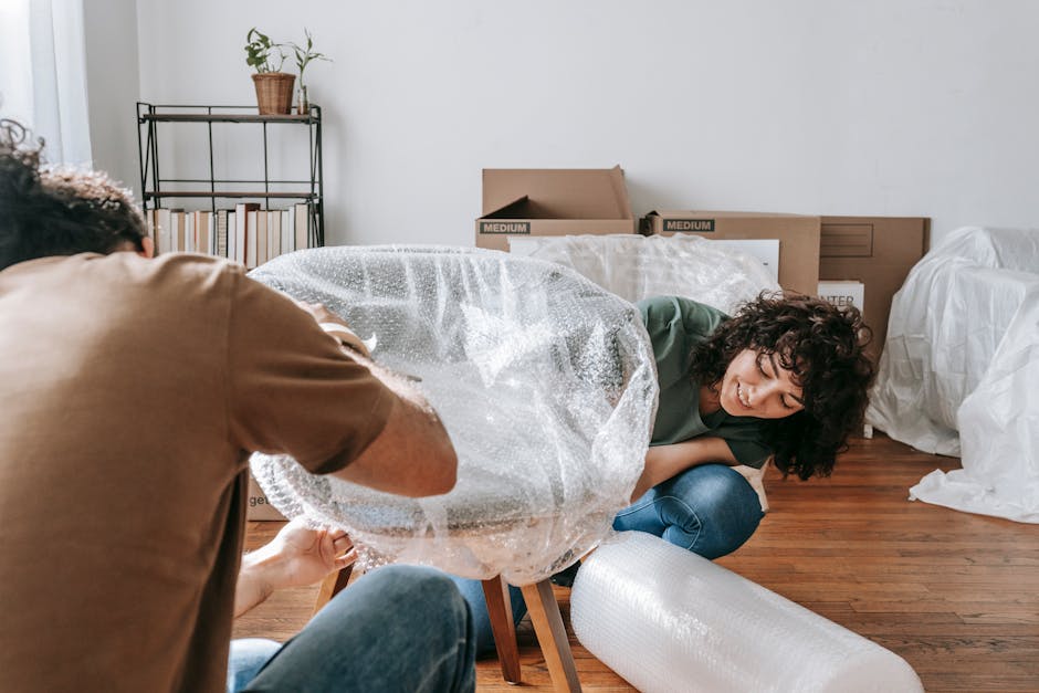 A woman with curly dark hair and a green top is seated on a hardwood floor inside a room, smiling as she helps with the packing process during a home relocation. She is holding a rolled-up piece of bubble wrap packaging material, which is being prepared for furniture or fragile items. A man wearing a brown T-shirt is partially visible in the foreground, actively wrapping a piece of valuable or delicate furniture with transparent bubble wrap. Behind them, cardboard boxes labeled 'Medium' are stacked on top of each other, and some are open, revealing packing materials. The room has white walls and is illuminated with natural light. In the background, there is a metal shelving unit with potted plants and neatly arranged books, alongside additional packing supplies and white protective sheets covering furnishings. The scene reflects the careful packing and loading process typical of house removals and furniture transport services provided by Man with Van Surbiton, supporting efficient and damage-free home relocations.