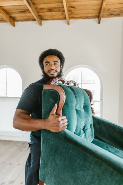 A man with dark curly hair and a beard is seen carrying a vintage-style armchair with a dark green velvet upholstery and ornate wooden detailing on the arms and backrest. He is inside a bright room with high ceilings featuring exposed wooden beams, large arched windows allowing natural light, and light-colored walls. The man is wearing a black t-shirt and black trousers, firmly grasping the chair’s wooden armrests as part of a home relocation process. The furniture is supported by his hands in preparation for moving, with additional packing materials such as protective blankets or padding not visible but implied. The scene depicts a furniture transport or packing and moving activity, typical of professional removals services provided by companies like Man with Van Surbiton, as they prepare to load the furniture onto a van for transportation. The environment and action suggest a careful and organized approach to furniture transport within a residential setting.