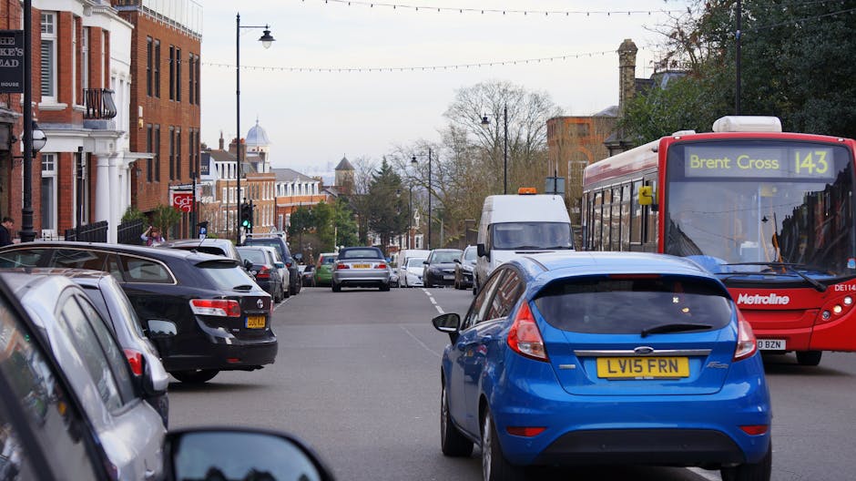 A busy urban street scene showing multiple parked cars along the curb, including a black van and a blue hatchback, with a white delivery van and a red Metroline bus on the road. The bus displays route information for Brent Cross on its electronic sign. Residential brick buildings with white window frames line the street, and there are street lamps, trees, and overhead power lines visible against an overcast sky. The scene captures moments of movement and parking, typical of a city environment engaged in home relocation or furniture transport preparations, with the surroundings indicating a residential area suitable for local house removals by [COMPANY_NAME].