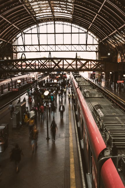 Inside a busy train station with a high, arched glass roof allowing natural light to illuminate the platform area, where numerous blurred passengers are walking, boarding, or leaving trains. On the right side, a red and black train is positioned along the platform with visible doors and windows. The platform surface shows tactile paving strips near the train for safety and accessibility. Overhead, structural metal beams support the roof, and the background reveals additional station features such as signs and lighting fixtures. This scene captures the movement and activity typical of a central rail hub, relevant to home relocation or moving logistics involving train travel and transportation.