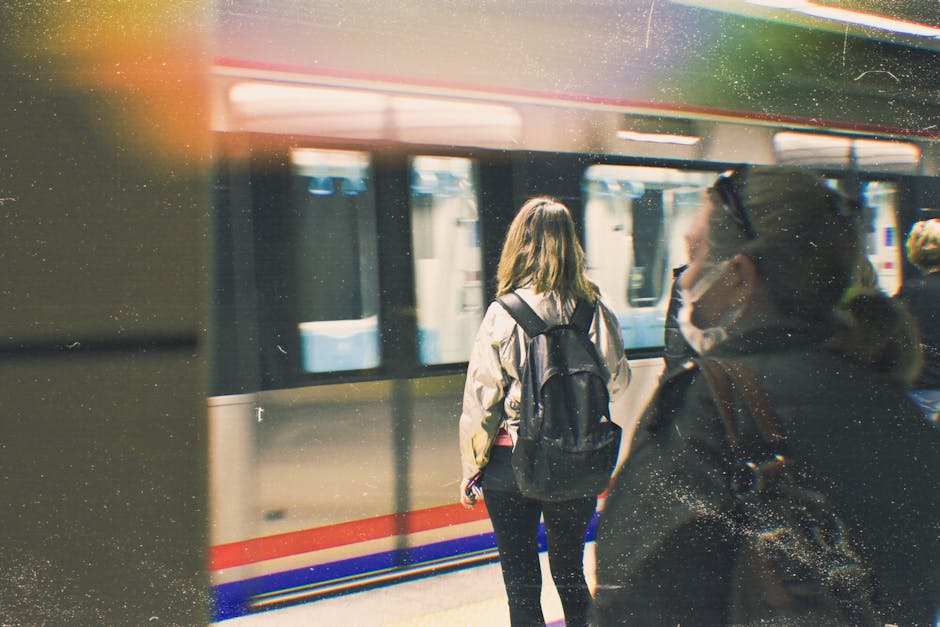 Two women standing on a platform at a train station, waiting for a train to arrive. The woman in the foreground is wearing a face mask, sunglasses, and a dark jacket, with a backpack on her shoulders. The woman in the background has long hair, wearing a light-colored jacket and a backpack, facing away from the camera toward the train. The train, with its sliding doors open, is visible behind them, showing the interior through the glass. The platform is well-lit with a modern ambiance, and the scene captures a typical scene of travel or home relocation preparations involving transportation. This setting relates to property moving and furniture transport services provided by companies such as Man with Van Surbiton, supporting the process of packing and logistics for house removals near Surbiton Station.