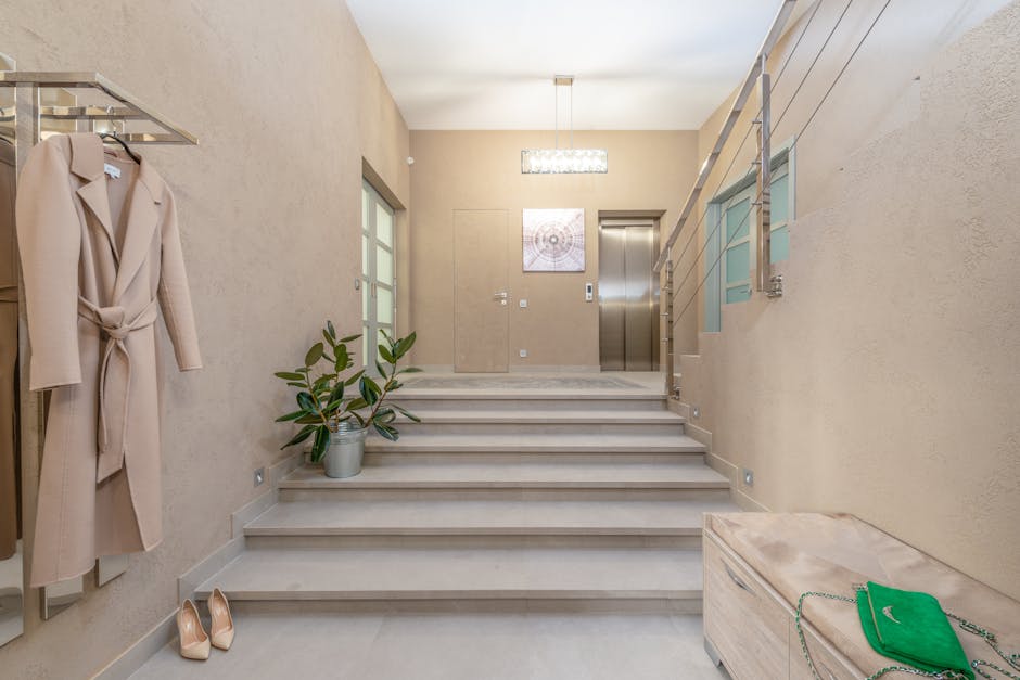 A spacious indoor hallway in a residential property featuring a staircase with beige steps and a metal railing leading to the upper floor. To the left, there is a clothing rack with a beige coat hanging, and a pair of nude high-heeled shoes on the floor beneath it. A green potted plant is positioned at the base of the staircase. The walls are painted in a neutral tone, and a modern rectangular light fixture hangs from the ceiling. At the top of the stairs, an elevator with closed doors is visible, alongside a door and a piece of abstract wall art. The right side of the image shows a section of the staircase railing with glass panels, and the area appears well-lit with natural and artificial lighting. This setting reflects the initial staging of a home moving process, with careful packing and accessibility considerations, aligned with professional removals services such as those by Man with Van Surbiton.