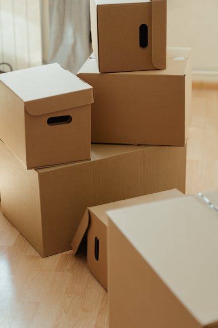 A collection of brown cardboard moving boxes of various sizes stacked and arranged on a light wooden floor inside a property, with some boxes featuring black plastic handle holes. The boxes are part of a home relocation process, prepared for packing and moving. In the background, there is a white wall and a partially visible window with curtains. The image depicts the initial stages of loading or organizing packing materials for a house removal, highlighting the careful use of packaging supplies typical of professional furniture transport and moving logistics by Man with Van Surbiton. The setting suggests an indoor environment with natural lighting, focusing on the orderly arrangement of boxes ready for transportation to a new location.
