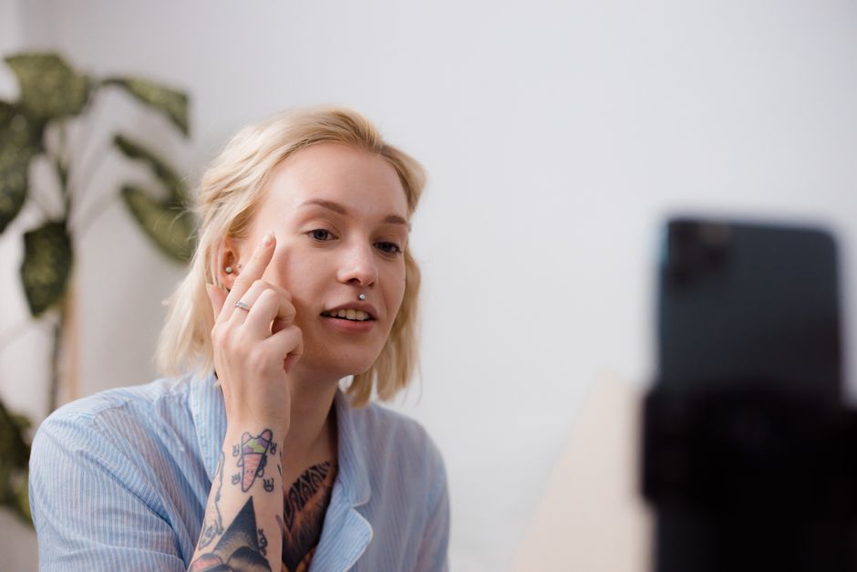 The image shows a young woman with short blonde hair and visible tattoos on her right arm, sitting indoors in a well-lit room with a white wall background and a potted plant with green leaves behind her. She is wearing a light blue, striped shirt and has a neutral expression on her face, with her right hand raised to her temple, making a gesture as if thinking or explaining. In front of her, a smartphone mounted on a tripod or stand captures her image, suggesting a video or photo recording. The setting appears casual and modern, with soft lighting illuminating her face. The scene may relate to home relocation or packing and moving activities, reflecting the process of communication or instruction often involved in furniture transport and moving logistics. This image is associated with Man with Van Surbiton, specialising in removals and house relocations in the Surbiton area, supporting the context of applying for moving permits in Surbiton (KT6).