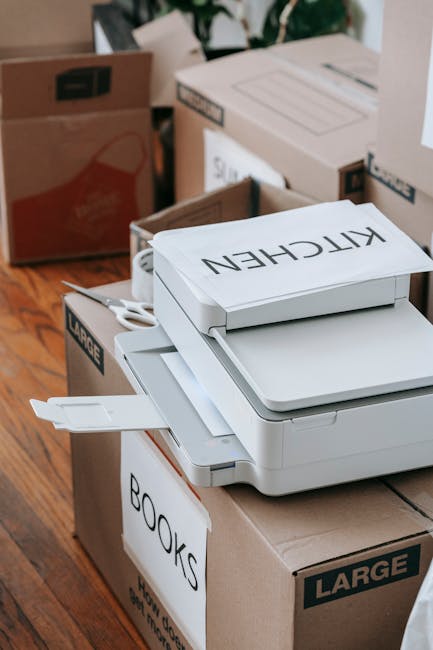 A stack of white office printers and printed labels, including one with the text 'KITCHEN', placed on top of large cardboard boxes labeled for house removals. The scene shows a home interior with wooden flooring and additional boxes in the background, some labeled for packing and moving purposes. The boxes are partially open, revealing packaging materials like cardboard and paper, and are arranged near a doorway or entryway indicating preparation for home relocation or furniture transport. The setting reflects the packing and loading process typical of professional removals services, as demonstrated by Man with Van Surbiton, with attention to careful handling of packing supplies and moving equipment visible in the scene.