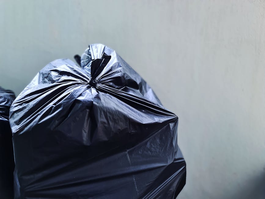 Close-up of several black plastic rubbish bags filled with waste, stacked on top of each other inside a property near an open doorway, with some bags appearing full and tightly tied at the top. The setting suggests a home relocation or packing process as part of a house move, with the bags likely prepared for disposal or rubbish collection in accordance with Surbiton Council rules. The background includes a plain, light-colored wall and a portion of the doorway, indicating an indoor environment during the packing and moving stage. The scene reflects the final stages of packing materials, such as waste bags, before transporting or disposing of them, with the potential involvement of a professional removals service like Man with Van Surbiton.