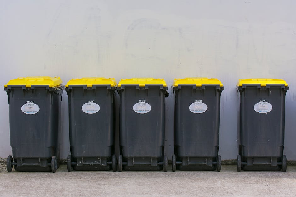 Five black wheelie bins with yellow lids lined up against a plain light grey wall in an outdoor setting, positioned on a paved surface. Each bin has a white label with black text, and they are placed side by side with minimal gap between them. The setting suggests an area designated for rubbish disposal as part of a home relocation process, with no visible items inside the bins. The image captures the bins from a frontal view, clearly showing their wheels and lids, and is associated with the moving and disposal regulations in Surbiton, as outlined by Surbiton Council. The professional removal company, Man with Van Surbiton, may utilize these bins for waste collection post-move, supporting efficient packing, disposal, and transportation logistics during the house removal process.